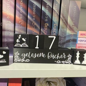 Puede incluir: Un bloque de madera negro con el texto "gelesene Bücher" en blanco. El bloque está sentado en un estante con otros bloques de madera negros que tienen números en ellos. Los números en los bloques son 17. Hay libros en el estante detrás de los bloques.