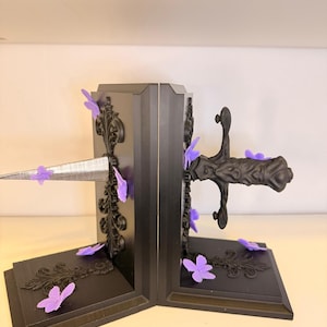 May include: Black bookends with ornate detailing and purple butterfly accents. A silver sword design pierces through the center. The bookends have a decorative, gothic style.