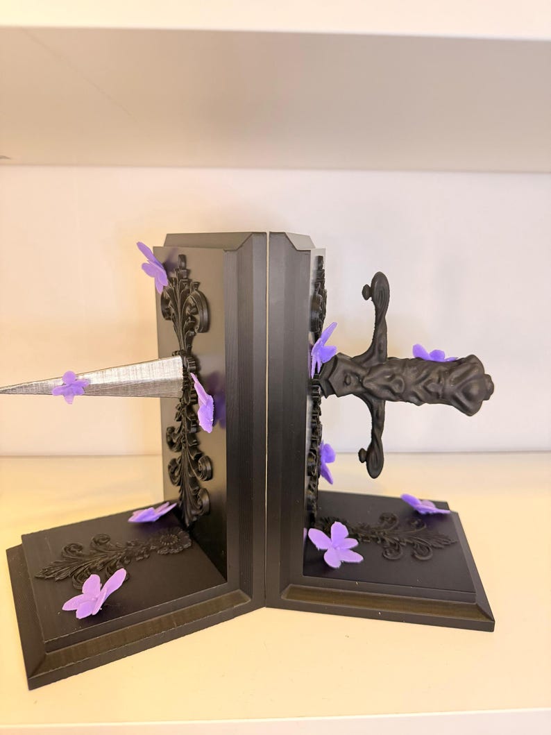 May include: Black bookends with ornate detailing and purple butterfly accents. A silver sword design pierces through the center. The bookends have a decorative, gothic style.