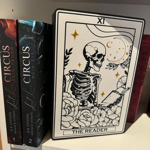 Könnte beinhalten: Eine rechteckige Dekotafel mit einer schwarz-weißen Tarotkarten-Illustration eines Skeletts, das ein Buch liest, umgeben von Blumen und einer Mondsichel. Das Design enthält goldene Akzente an Sternen und Mond sowie den Schriftzug THE READER.