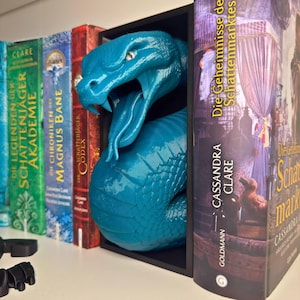 Könnte beinhalten: Ein tealblauer Schlangen-Buchstütze mit einem bedrohlichen Ausdruck, positioniert zwischen einer Reihe von Büchern mit deutschen Titeln. Der Buchstütze ist aus einem glatten, glänzenden Material gefertigt und scheint aus Kunststoff zu sein.