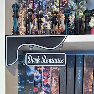 May include: A bookshelf with decorative chess pieces and books. The shelf features a sign that reads "Dark Romance". The chess pieces are black with gold, blue, and green accents. The books have floral and skull designs.