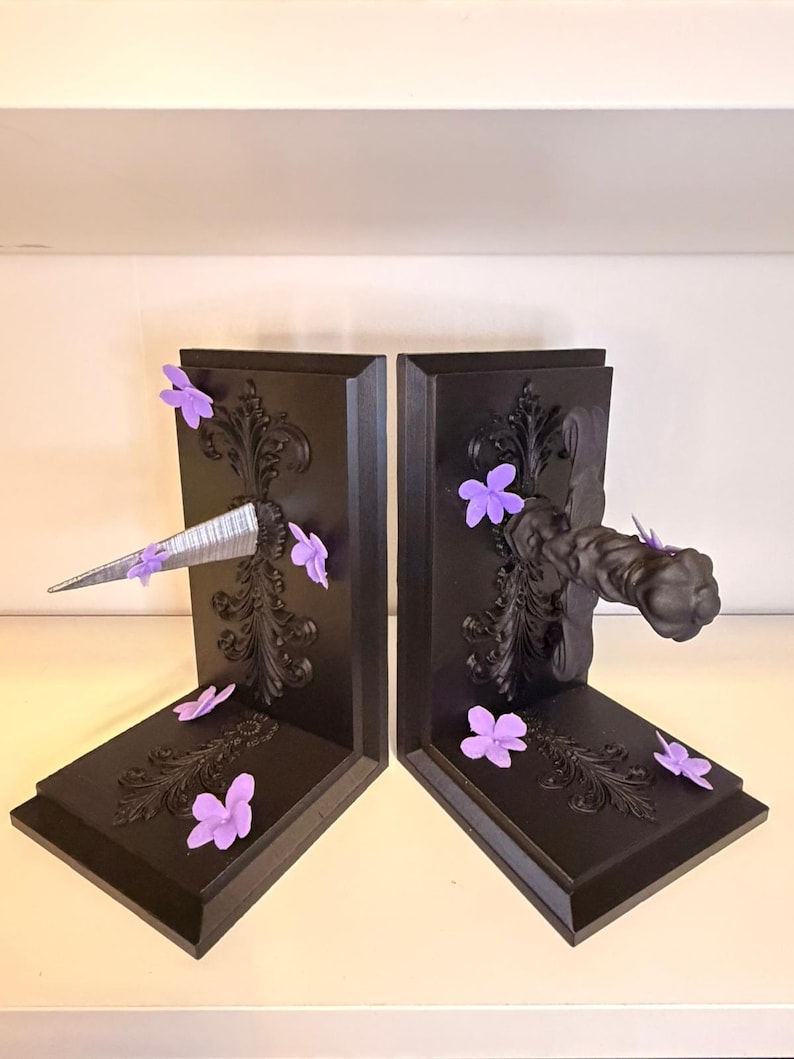 May include: A pair of black bookends with ornate scrollwork and purple flower accents. One bookend features a silver, metallic spike, while the other has a decorative handle. The bookends are designed to hold books upright.