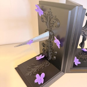 May include: Black bookends with ornate scrollwork and purple flower accents. The bookends have a decorative design and a silver, triangular element. The bookends are on a white surface.