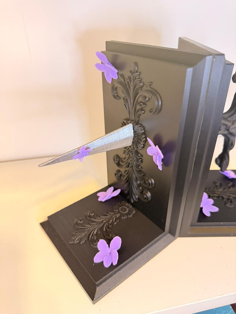 May include: Black bookends with ornate scrollwork and purple flower accents. The bookends have a decorative design and a silver, triangular element. The bookends are on a white surface.