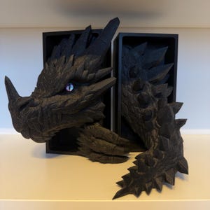 Könnte beinhalten: Schwarze Drachen-Buchstützen mit detailliertem, strukturiertem Design. Der Kopf und der Schwanz des Drachen sind auf beiden Seiten der Bücher positioniert, mit einem einzelnen, irisierenden Auge. Die Buchstützen bestehen aus dunklem Material und sind so konzipiert, dass sie Bücher aufrecht halten.