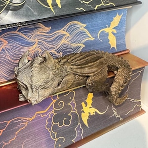 Op de afbeelding: Een bronzen drakenbeeldje rust op een stapel decoratieve boeken. De boeken hebben gouden draken- en wolkenontwerpen op hun ruggen. De boeken zijn in tinten blauw, paars en zwart.