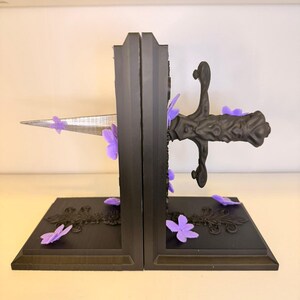 May include: Black bookends with a sword design. The bookends are decorated with purple flower accents. The sword appears to be piercing through the bookends. The base of the bookends have a decorative floral design.