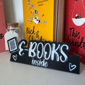 Könnte beinhalten: Ein schwarzes Schild mit den Worten "E-BOOKS inside" in weißer Schrift, verziert mit weißen Herzen. Das Schild steht vor zwei Büchern mit farbenfrohen Einbänden. Ein Buch ist gelb mit dem Titel "Nick & Charlie".