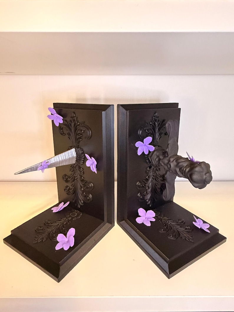 May include: A pair of black bookends with ornate scrollwork and purple flower accents. One bookend features a silver, pointed object, while the other has a decorative handle. The bookends are designed to hold books upright.