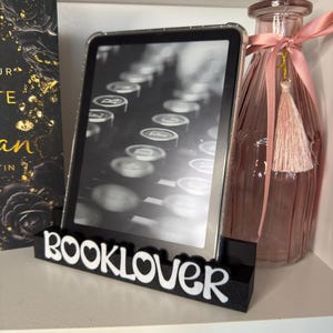 Può includere: Un supporto nero e bianco "BOOKLOVER" tiene un e-reader che mostra un'immagine di macchina da scrivere. Una bottiglia di vetro rosa con nappa e nastro è a destra. Un libro con lettere dorate è a sinistra.