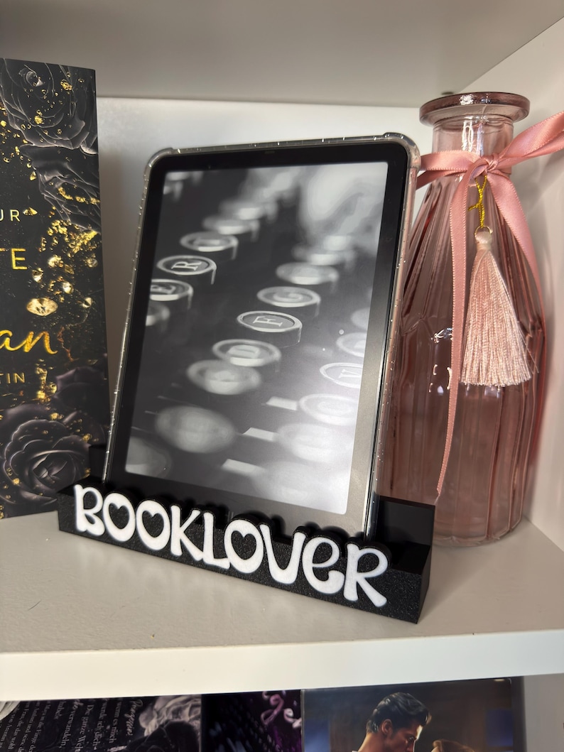 Puede incluir: Un soporte negro y blanco "BOOKLOVER" sostiene un lector electr&oacute;nico que muestra una imagen de m&aacute;quina de escribir. Una botella de vidrio rosa con borla y cinta est&aacute; a la derecha. Un libro con letras doradas est&aacute; a la izquierda.