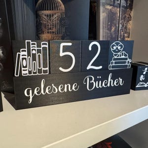 Könnte beinhalten: Schwarze Holzblöcke mit weißem Text und Illustrationen. Die Blöcke buchstabieren "52" und zeigen einen Stapel Bücher und einen Bücherwurm. Der untere Block lautet "gelesene Bücher".