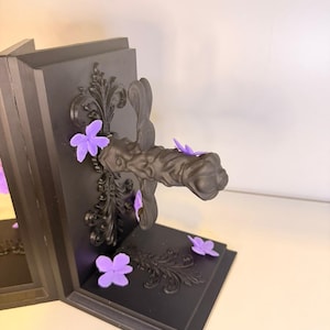 May include: Black bookends with ornate scrollwork and purple flower accents. The bookends have a dark, matte finish and a decorative handle. The flowers are scattered around the base and add a pop of color to the design.
