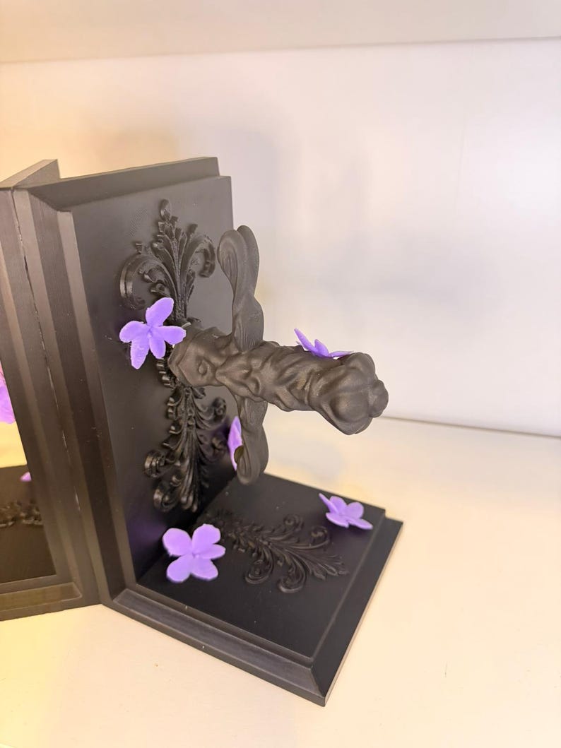 May include: Black bookends with ornate scrollwork and purple flower accents. The bookends have a dark, matte finish and a decorative handle. The flowers are scattered around the base and add a pop of color to the design.