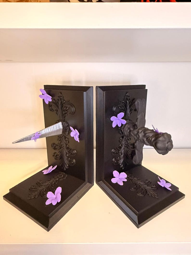 May include: A pair of black bookends with ornate scrollwork and purple flower accents. One bookend features a silver, metallic spike, while the other has a black, fist-shaped handle. Decorative and functional.