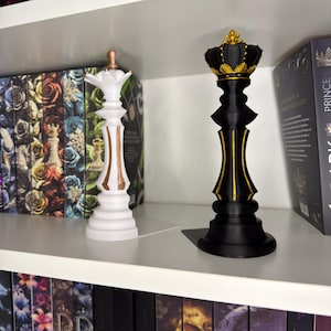Könnte beinhalten: Ein Paar dekorative Buchstützen in Form von Schachfiguren. Die weiße Buchstütze ist eine Dame mit einer goldenen Krone und Verzierungen. Die schwarze Buchstütze ist ein König mit einer goldenen Krone und Verzierungen.