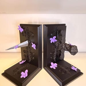 May include: Pair of black bookends with ornate scrollwork and purple flower accents. One bookend features a silver, pointed object, while the other has a decorative handle. The bookends are designed to hold books upright.
