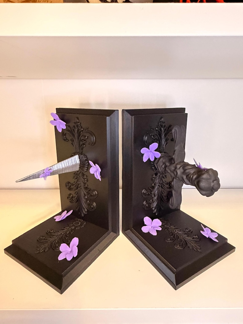 May include: Pair of black bookends with ornate scrollwork and purple flower accents. One bookend features a silver, pointed object, while the other has a decorative handle. The bookends are designed to hold books upright.