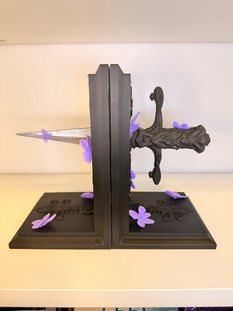 May include: Black bookends with a sword design, accented with purple flower embellishments. The bookends have a rectangular base with decorative scrollwork. The sword blade is silver, and the handle is black.