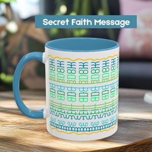 Op de afbeelding: Witte keramische mok met een blauwe binnenkant en handvat. De mok heeft een herhalend patroon van groene en blauwe tekst en decoratieve lijnen. De tekst "Secret Faith Message" wordt weergegeven in een blauwe banner bovenaan.