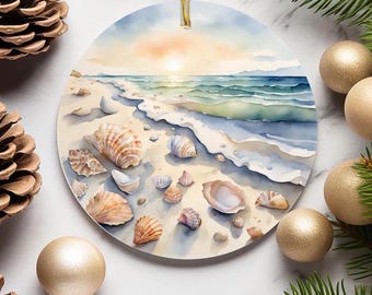 Puede incluir: Adorno navideño redondo con una escena de playa en acuarela, con conchas marinas, arena y olas bajo un atardecer. El adorno tiene una cuerda dorada para colgar. Piñas y adornos dorados rodean el adorno.