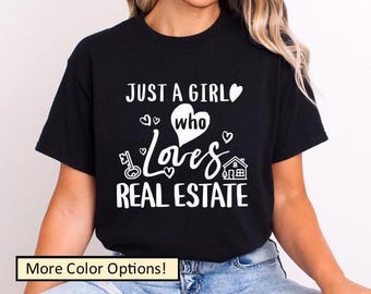 May include: Black t-shirt with white text that reads "JUST A GIRL who Loves REAL ESTATE." The design includes a heart, key, and house graphic. The shirt is a crew neck and has short sleeves.