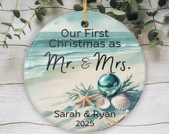 Puede incluir: Adorno navideño redondo con un diseño de escena de playa. El adorno presenta el texto "Our First Christmas as Mr. & Mrs." junto con los nombres "Sarah & Ryan" y el año "2025". Incluye una bola turquesa, estrellas de mar y conchas.
