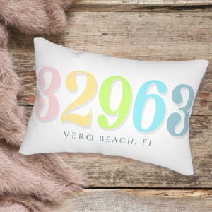 Könnte beinhalten: Ein weißes Kissen mit den Zahlen 32963 in Pastellrosa, Gelb, Grün, Blau und Grau. Der Text "VERO BEACH, FL" ist unter den Zahlen gedruckt.