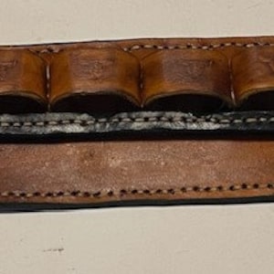 May include: A brown leather belt with six loops for holding ammunition. The belt has a black leather strip across the top and a decorative circle on the left side.