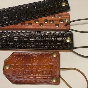 May include: Four leather and wood items. Two black leather pieces with embossed text that reads "SIDEWINDER" and "GOD GAVE ME SIDEWINDER". One wooden piece with holes. One brown leather piece with the text "SLINGING SMOKE".