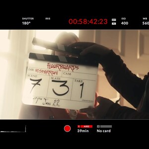 Peut inclure: Un clap de cinéma avec le titre "FLOORBOARDS" et des informations de scène, de prise et de date. Le clap est tenu par une personne, avec l'affichage du timecode en arrière-plan. La date "17th Jan 22" est également visible.
