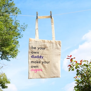 Puede incluir: Una bolsa de tela blanca con el texto "be your own daddy. make your own sugar" impreso en ella. La bolsa está colgada de un tendedero contra un cielo azul.