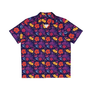 May include: A short-sleeved button-up shirt with a colorful, all-over print. The print features a repeating pattern of gold bars, crescent moons, and stylized flowers. The shirt is a dark blue color.