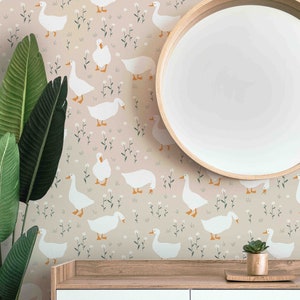 Op de afbeelding: Een lichtroze behang met een herhalend patroon van witte ganzen en kleine witte bloemen. Het behang bevindt zich achter een houten kast met twee witte lades. Een ronde houten spiegel hangt boven de kast.