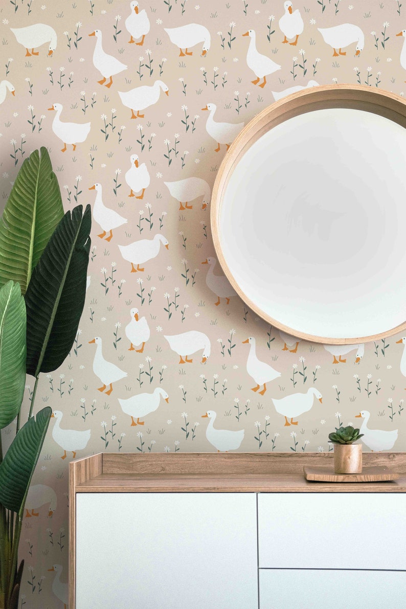 Op de afbeelding: Een lichtroze behang met een herhalend patroon van witte ganzen en kleine witte bloemen. Het behang bevindt zich achter een houten kast met twee witte lades. Een ronde houten spiegel hangt boven de kast.