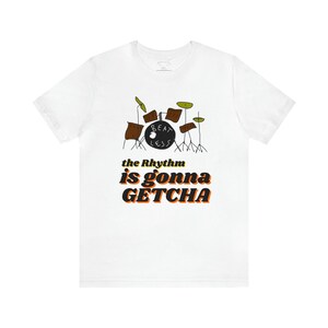 May include: White t-shirt featuring a cartoon drum set illustration in brown, green, and yellow. The text "the Rhythm is gonna GETCHA" is printed below the drum set in orange and black.