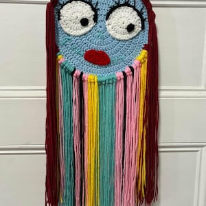 Può includere: Una decorazione da parete all'uncinetto fatta a mano con un volto circolare azzurro, grandi occhi bianchi, pupille nere, labbra rosse e dettagli cuciti. L'oggetto presenta lunghi capelli in lana rosso scuro e frange di fili multicolori rosa, gialli, turchesi e neri.