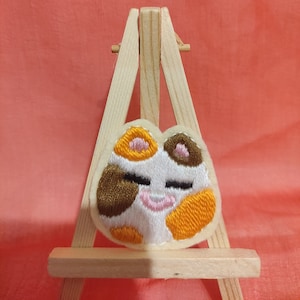 Peut inclure: Un patch brodé blanc, marron et orange représentant un chat les yeux fermés. Le patch est présenté sur un petit chevalet en bois.