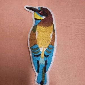 Peut inclure: Écusson brodé d'un oiseau coloré avec un long bec noir. L'oiseau présente une poitrine jaune, des ailes bleues et une tête brune. L'écusson est bordé de coutures blanches et est posé sur un fond rose.