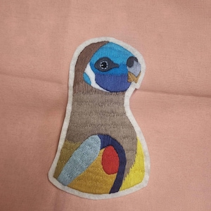 Könnte beinhalten: Gestickter Aufnäher mit einem stilisierten Vogeldesign. Der Vogel hat einen blauen Kopf, einen grauen Körper und eine gelb-rote Brust. Der Aufnäher ist mit einem weißen Rand umrandet und vor einem rosa Hintergrund platziert.