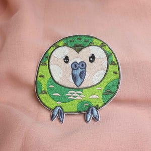 Può includere: Toppa ricamata con un design di pappagallo dei cartoni animati. La toppa è verde e bianca con accenti blu. Il pappagallo ha una forma del corpo rotonda ed è ambientato su uno sfondo rosa.