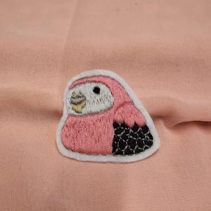 May include: An embroidered patch of a bird, featuring pink and black colours with a white outline.