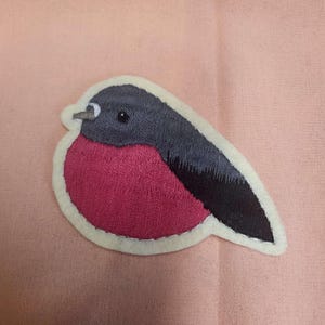 Peut inclure: Un patch brodé en feutre représentant un rouge-gorge avec une tête noire, des ailes grises et une poitrine rouge.