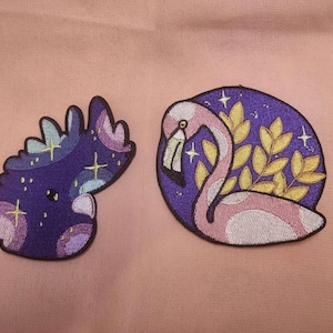 May include: Two embroidered iron-on patches. One patch features a purple and pink stylized creature with stars and swirls. The other patch features a pink flamingo with gold leaves on a purple background.