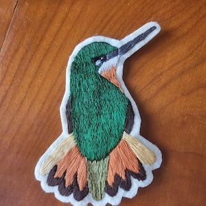 May include: Embroidered hummingbird patch with a white border. The bird features green, orange, brown, and tan thread. The hummingbird's beak is black, and the body is green. The patch is on a wooden surface.