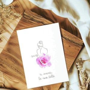 Può includere: Una carta bianca con un disegno a tratto di una silhouette femminile, una rosa rosa e il testo italiano "ti meriti le cose belle". La carta è su una superficie di legno con un tessuto bianco.