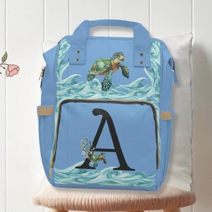 May include: A light blue backpack with a sea turtle design and the letter "A" on the front. The backpack is sitting on a wooden stool with a light brown cushion.