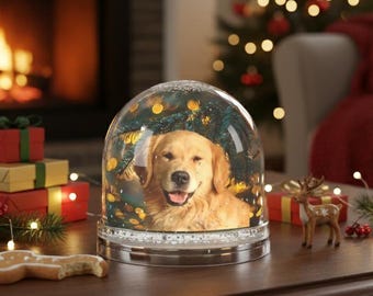 Op de afbeelding: Een sneeuwbol met een Golden Retriever hond, tegen een achtergrond van een kerstboom. De bol staat op een donkere houten tafel met feestelijke decoraties, een peperkoek en een open haard op de achtergrond.