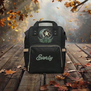 May include: Black diaper bag with a moon and star design and the name "Everly" embroidered on the front. The bag is sitting on a wooden surface with autumn leaves scattered around it.